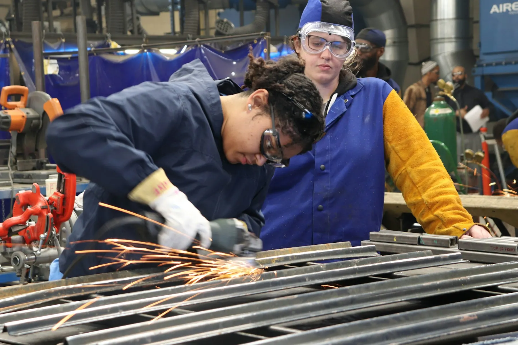 Two trainees at a workbench in a welding shop, one grinding metal while the other looks on